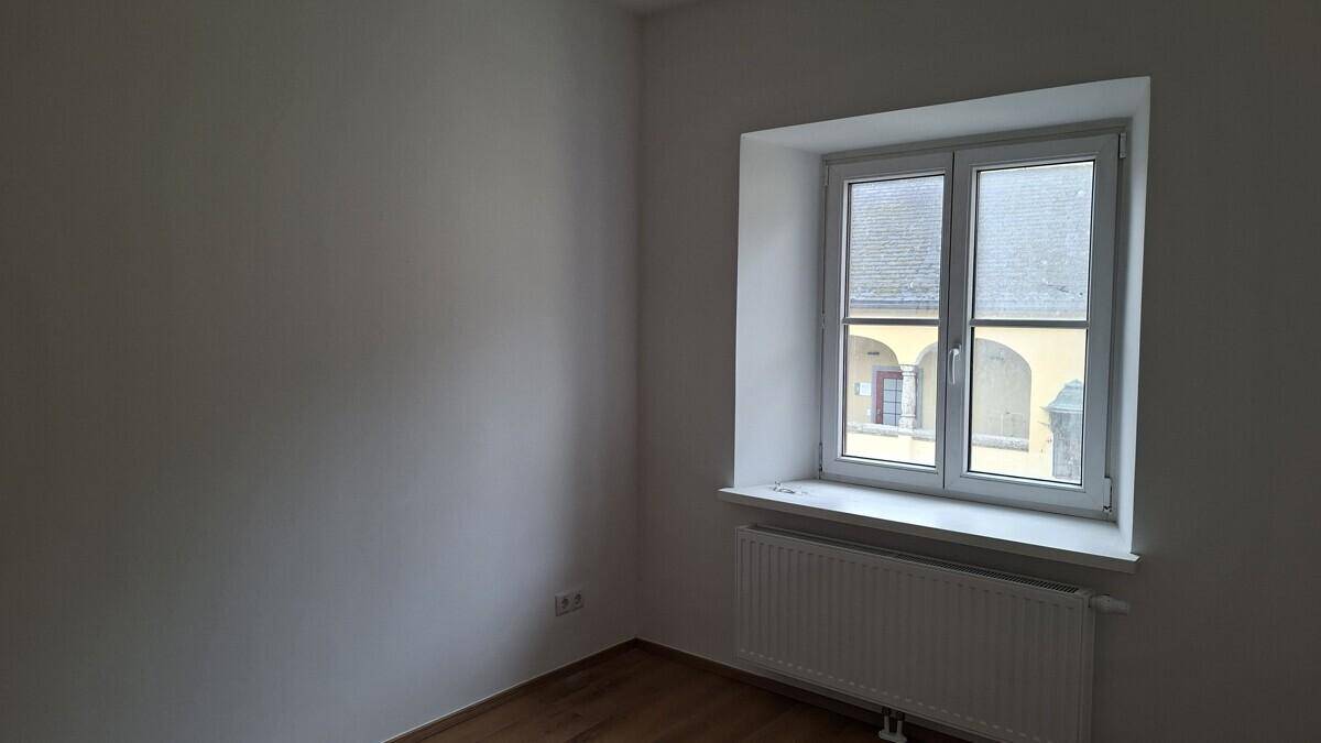 Leeres Zimmer mit Fenster und Heizkörper, bietet einen Blick auf einen Innenhof.