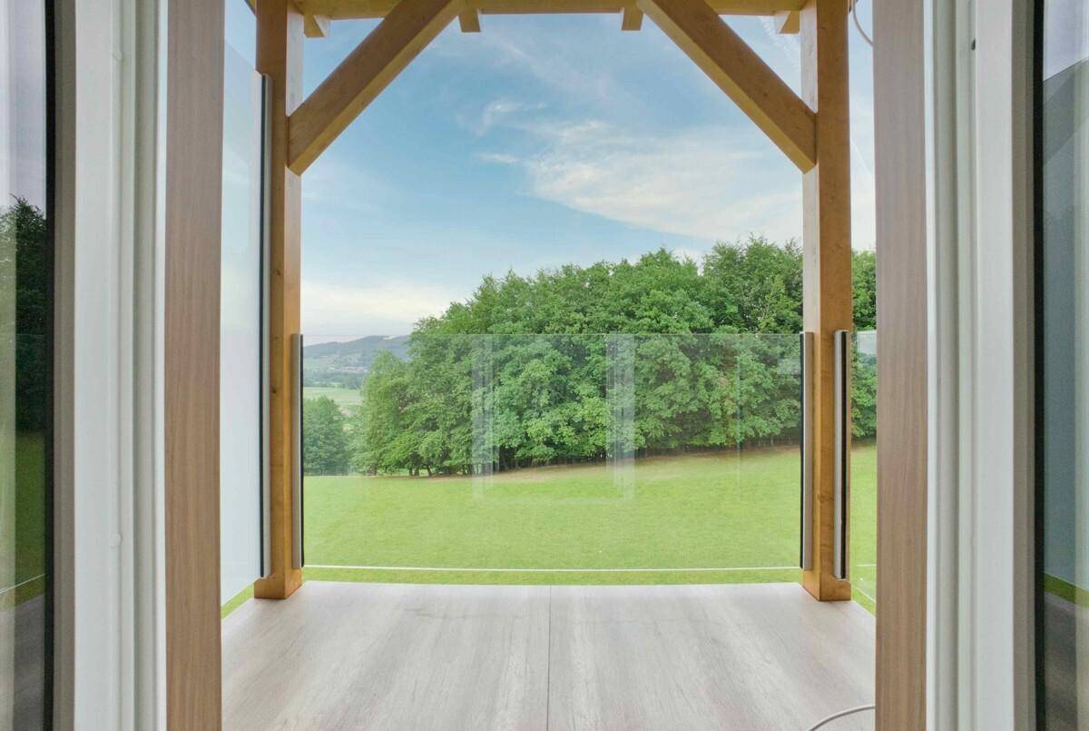 Balkon mit Holzkonstruktion und Glasgeländer, der einen weiten Blick ins Grüne bietet.