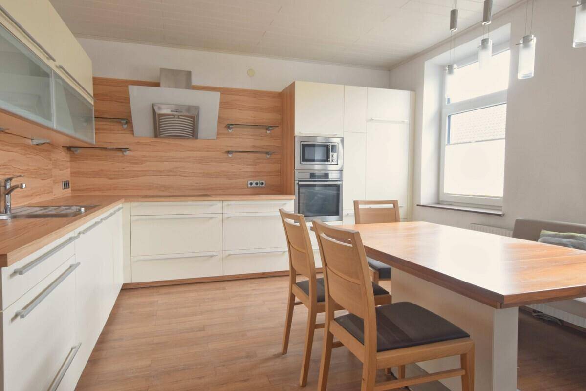 Moderne Wohnküche mit weißen Schränken, Holzakzenten und Essbereich am Fenster.
