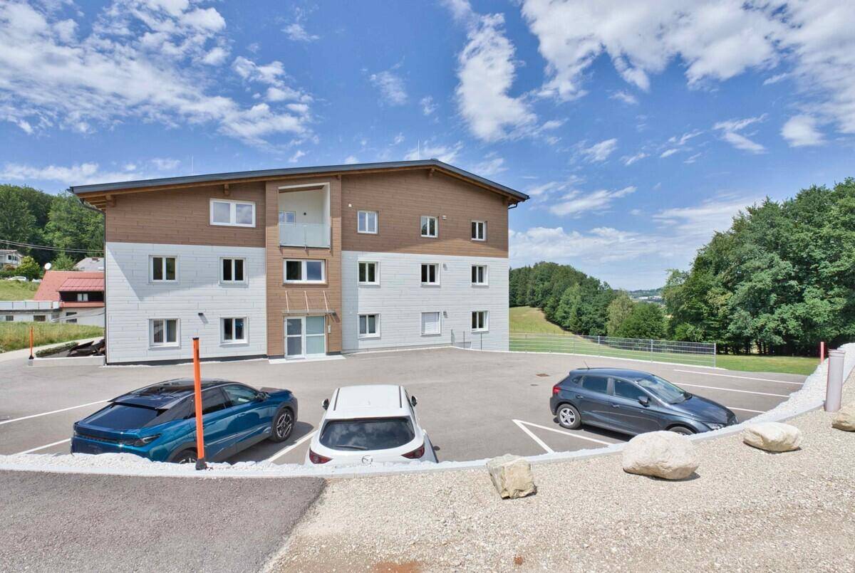 Modernes Mehrfamilienhaus mit Holz- und Putzfassade, umgeben von grüner Landschaft und Parkplätzen.