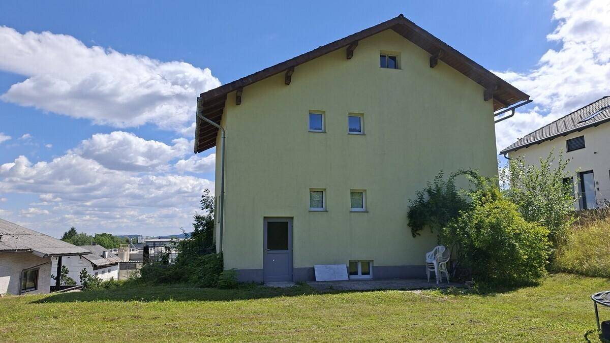 Rückseite des Hauses mit Rasenfläche und kleiner Terrasse, umgeben von Bäumen und Sträuchern.