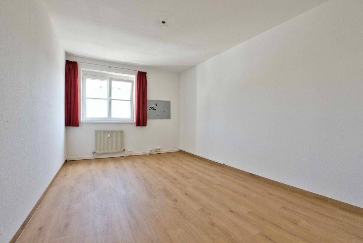Unmöblierter Raum mit Laminatboden, einem Fenster mit roten Vorhängen und einer weißen Wandgestaltung.