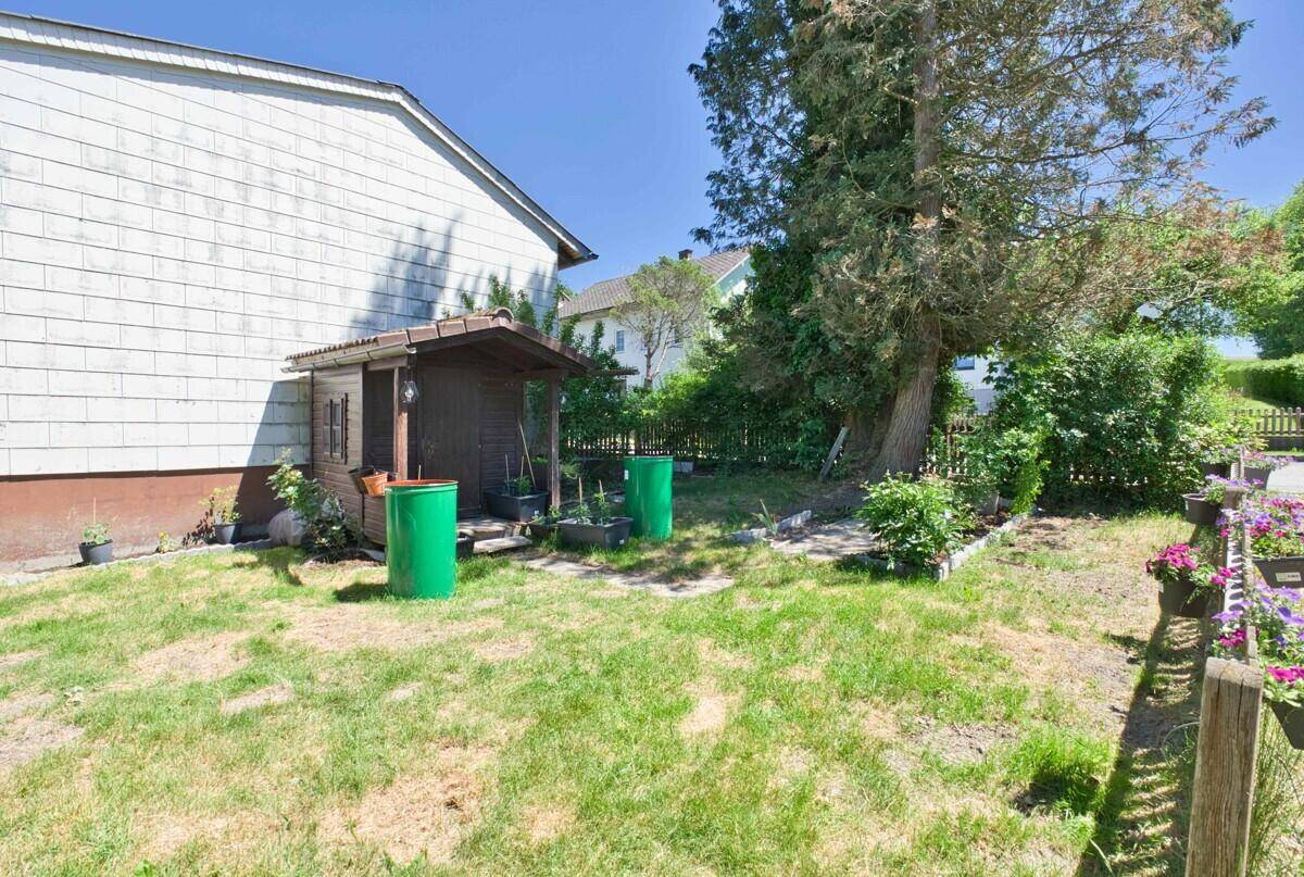 Kleiner Gartenbereich neben dem Haus mit einem Holzschuppen und grünen Tonnen unter blauem Himmel.