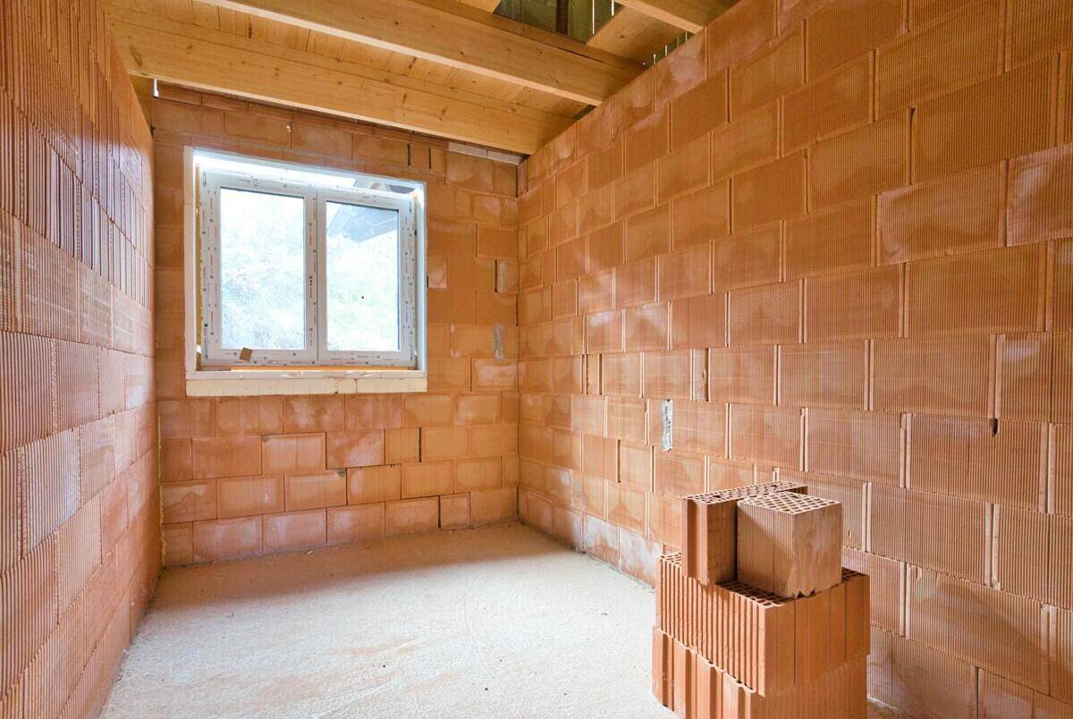 Kleinerer Raum im Rohbau mit Ziegelwänden, einem Fenster und sichtbaren Holzdeckenbalken.