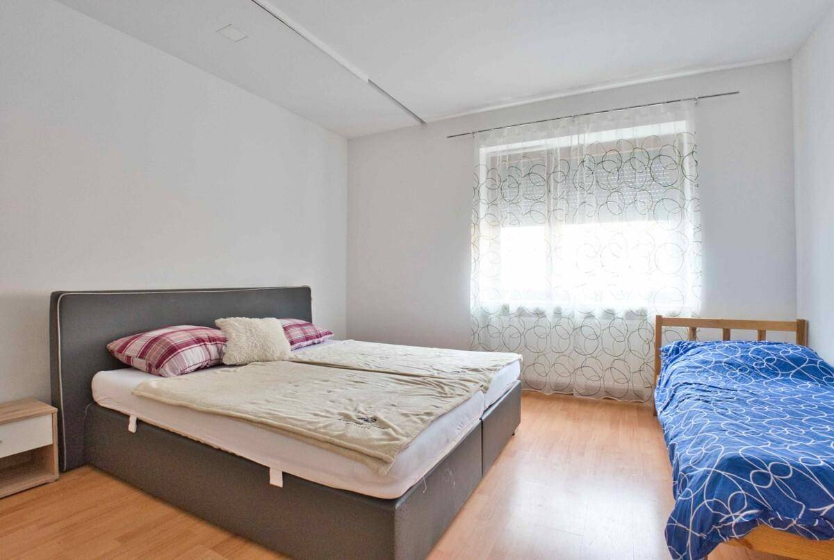 Schlafzimmer mit Doppelbett, Einzelbett und hellem Laminatboden, gut beleuchtet durch ein Fenster.