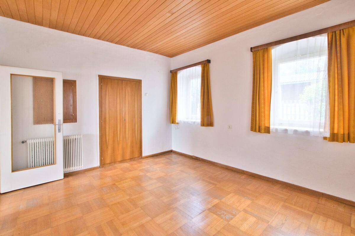 Schlafzimmer mit Holzparkettboden, Holzdecke und zwei Fenstern mit gelben Vorhängen.