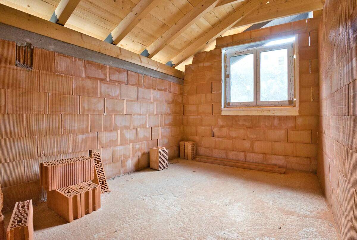 Raum im Rohbau mit schräger Decke, Ziegelwänden, einem Fenster und sichtbaren Holzbalken im Dachgeschoss.