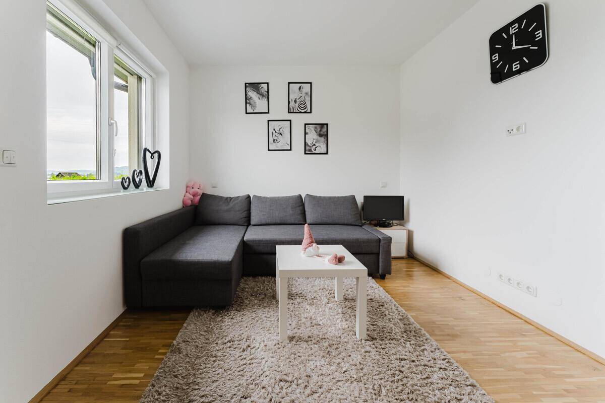 Freundliches Zimmer mit Holzboden, Sofa und moderner Wanduhr, vielseitig nutzbar.