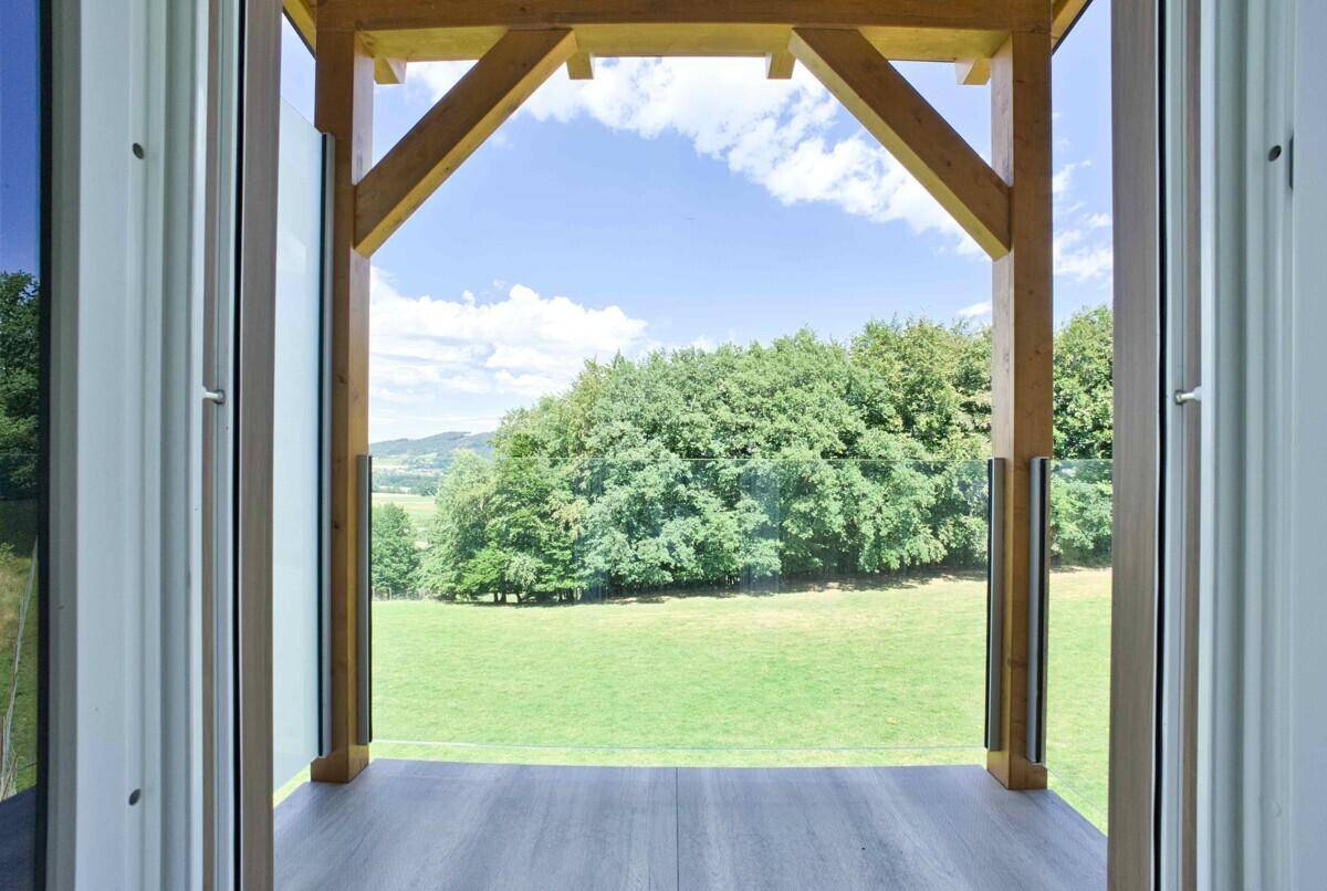 Heller Balkon mit Holzbalken und weitem Blick über grüne Landschaft und bewaldete Hügel unter blauem Himmel.