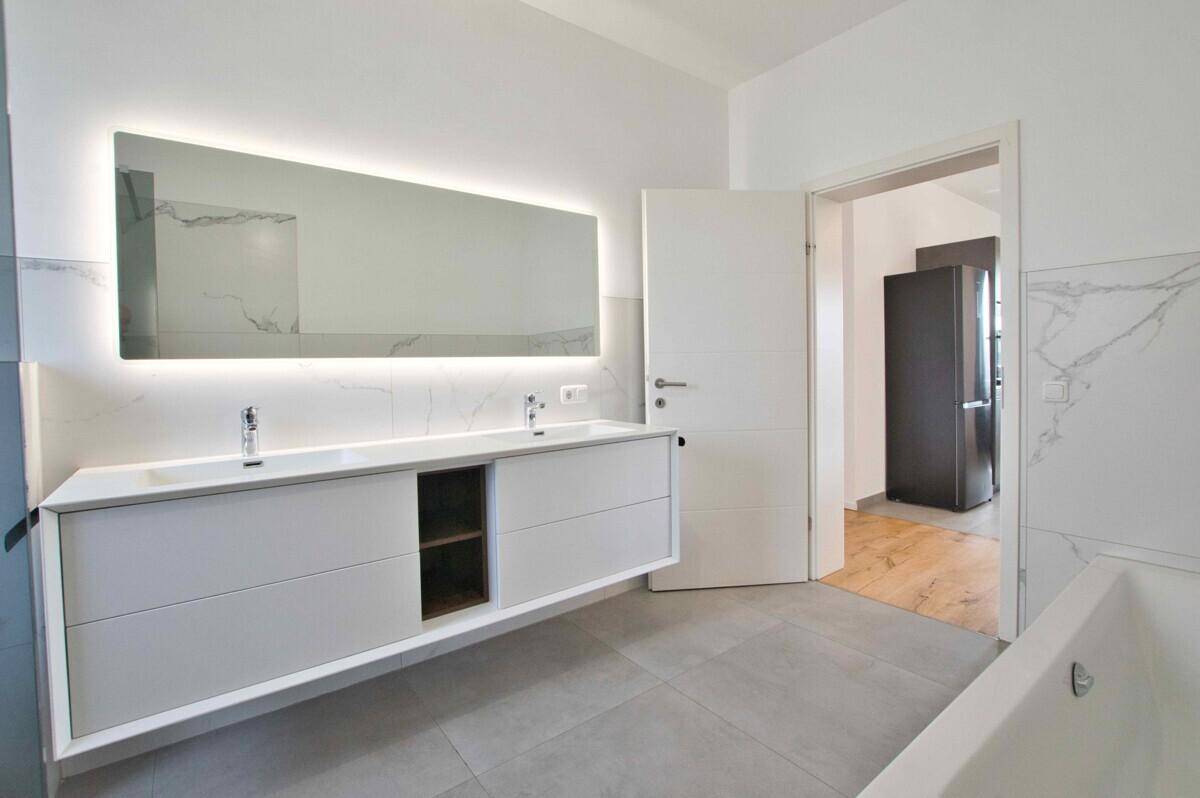 Elegantes Badezimmer mit beleuchtetem Spiegel, Doppelwaschbecken und Zugang zu einem weiteren Raum.