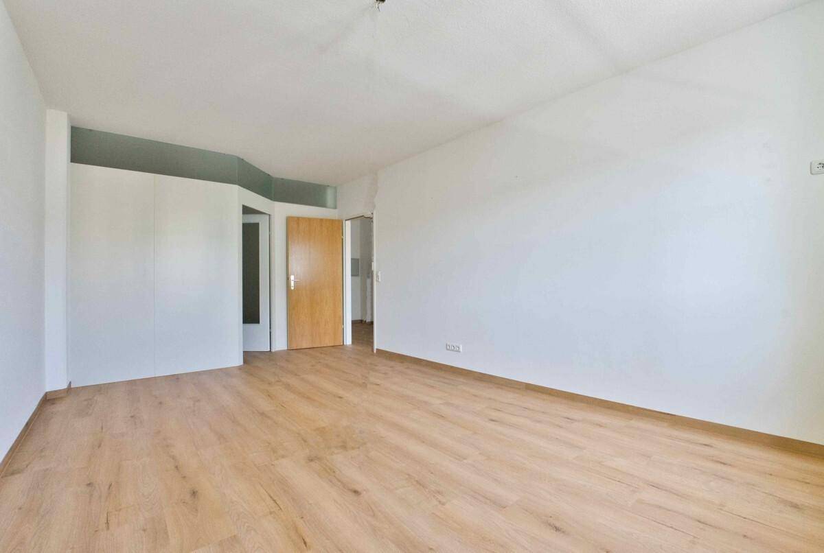 Geräumiger, leerer Raum mit hellem Laminatboden, weißen Wänden und mehreren Türen.