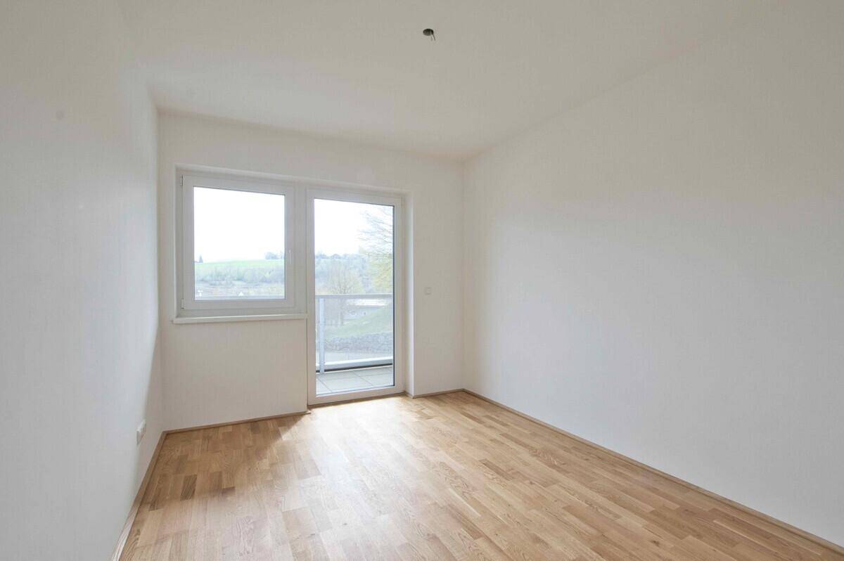 Leeres Schlafzimmer mit Holzparkettboden und Zugang zu einem kleinen Balkon.