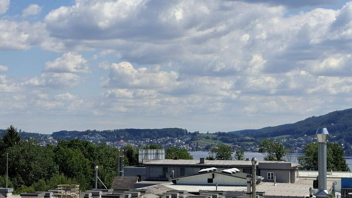 Panoramablick über Dächer einer Stadt am Seeufer mit bewaldeten Hügeln im Hintergrund.