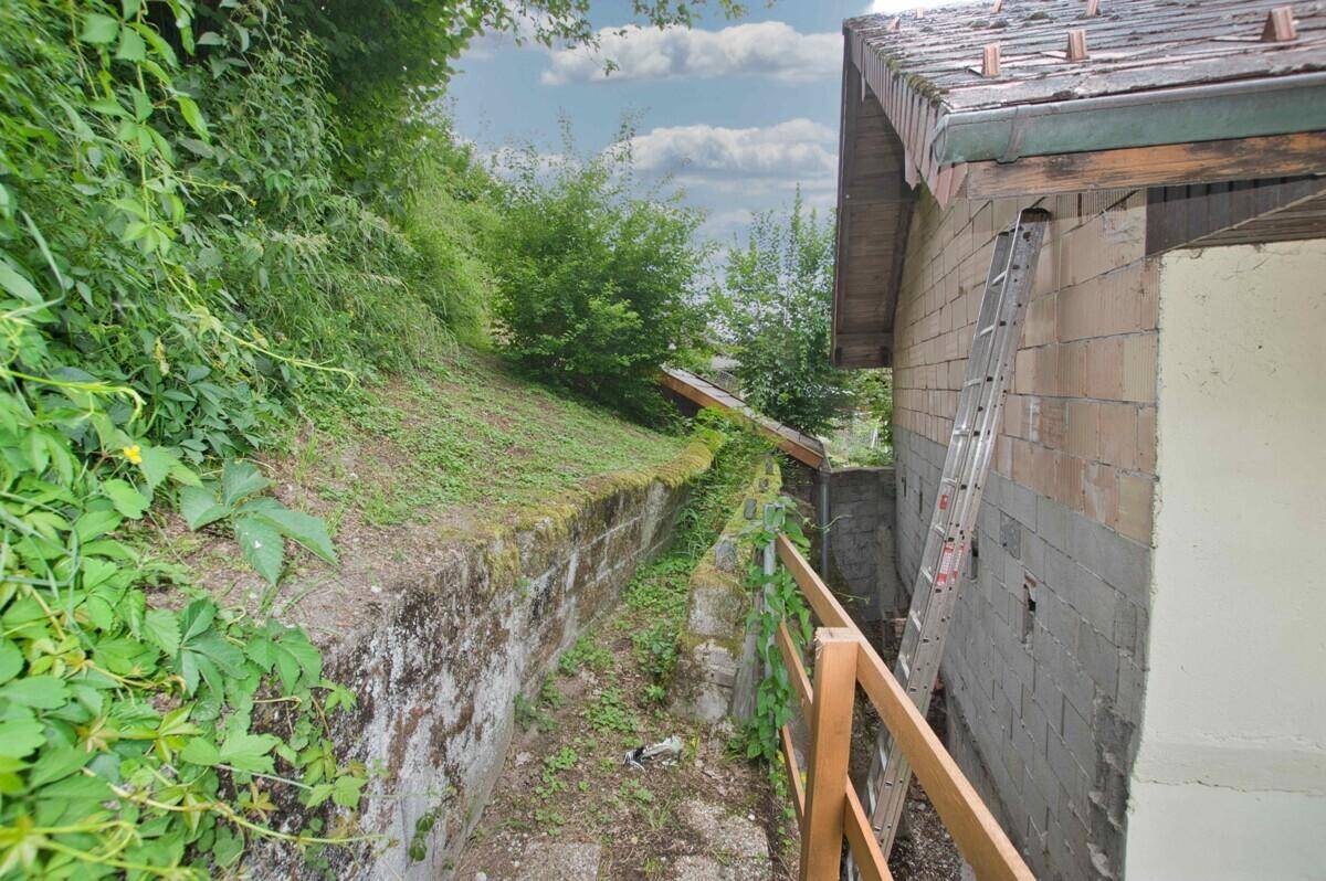 Schmaler, bewachsener Weg entlang einer Hauswand mit Leiter und Hanglage.