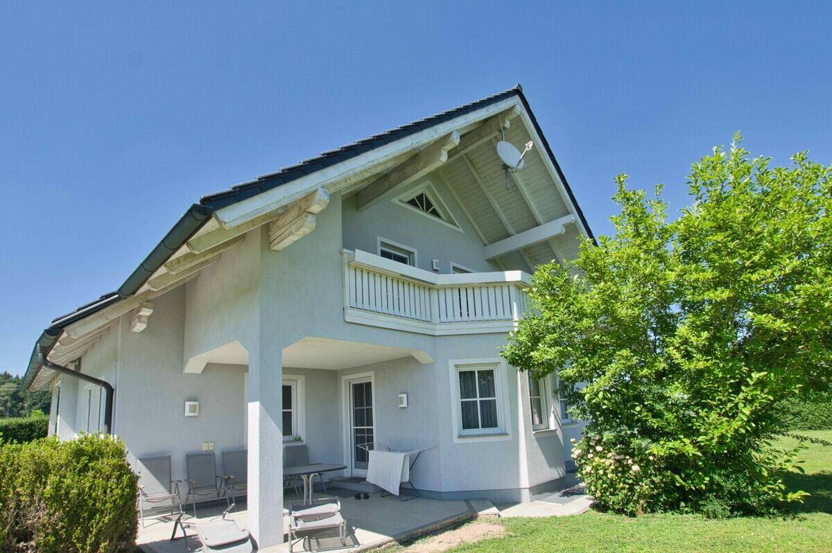 Überdachte Terrasse mit Sitzgelegenheiten und Balkon darüber, umgeben von Grün.