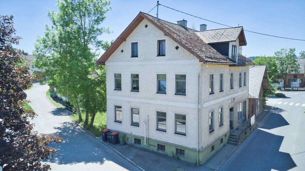 Mehrfamilienhaus mit heller Fassade und traditionellem Dach in ländlicher Umgebung unter blauem Himmel.