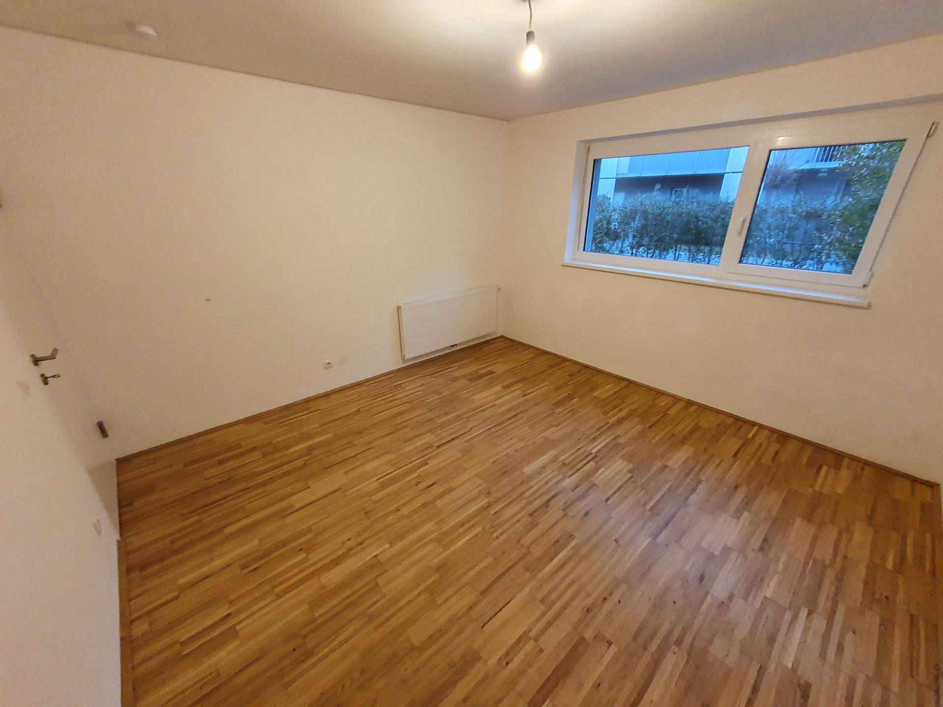 Helles Schlafzimmer mit Parkettboden und großem Fenster, das viel Tageslicht hereinlässt.