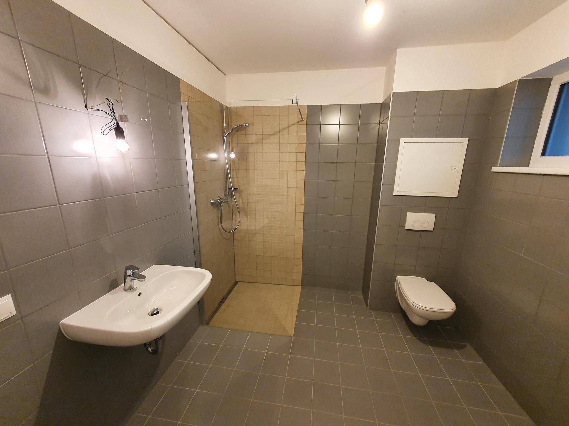 Modernes Badezimmer mit grauen Fliesen, bodengleicher Dusche und wandhängendem WC.