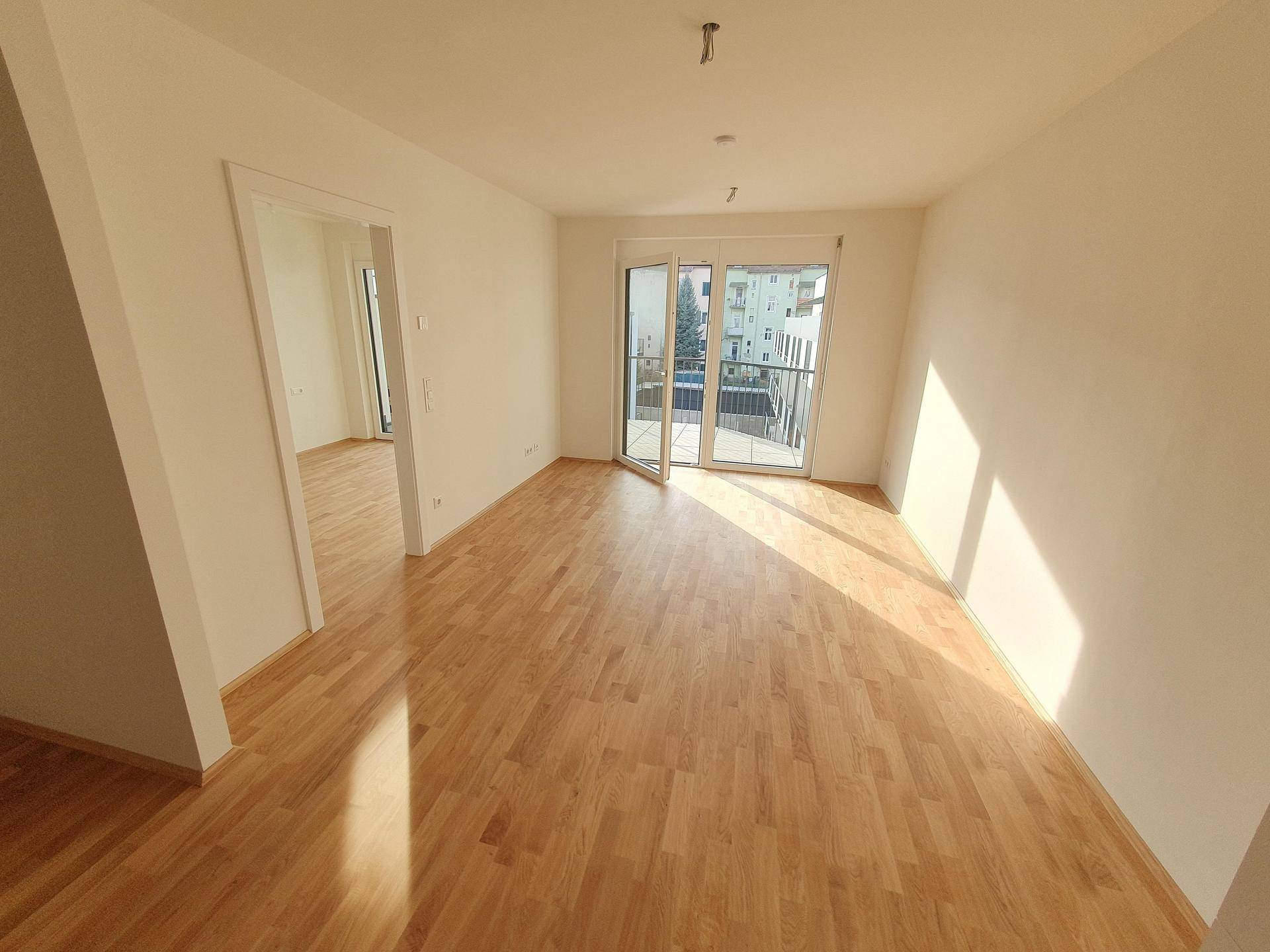 Heller Wohnbereich mit Holzboden, großen Fenstern und Zugang zu einem kleinen Balkon.