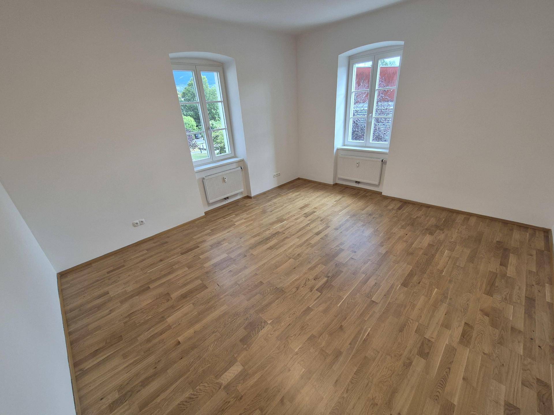 Heller Raum mit neuem Holzparkettboden und zwei Fenstern, die viel Tageslicht hereinlassen.