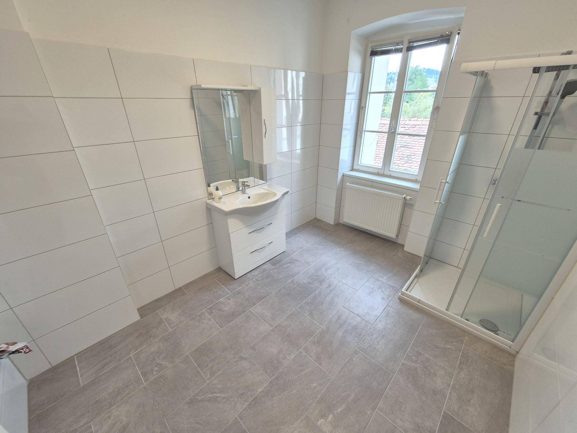 Modernes Badezimmer mit weißen Fliesen, Duschkabine, Waschbecken und Fenster für Tageslicht.
