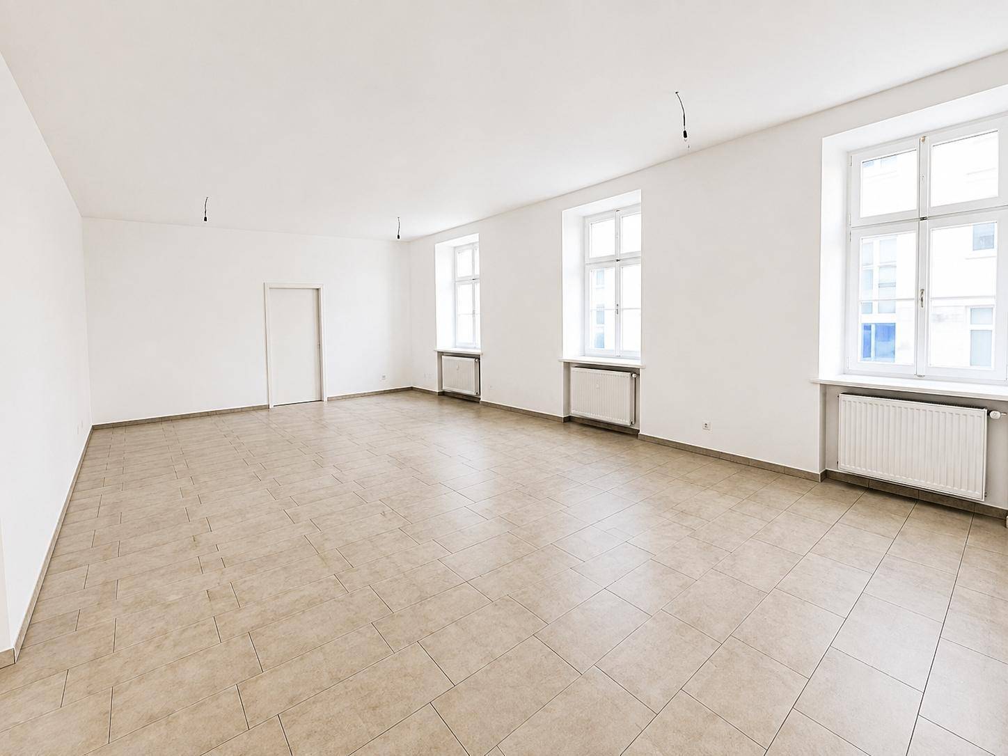 Heller, leerer Raum mit großen Fenstern, beigefarbenem Fliesenboden und Heizkörpern unter den Fenstern.