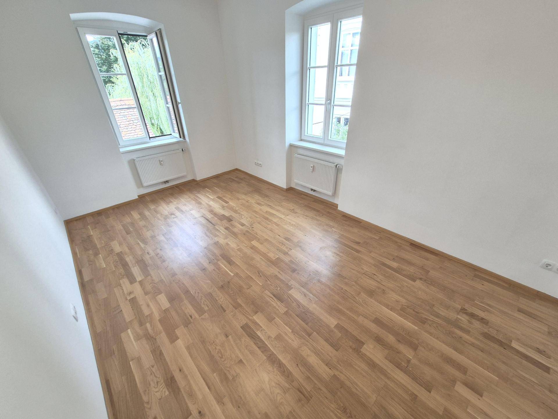 Geräumiger, lichtdurchfluteter Raum mit elegantem Holzparkett und Blick ins Grüne.