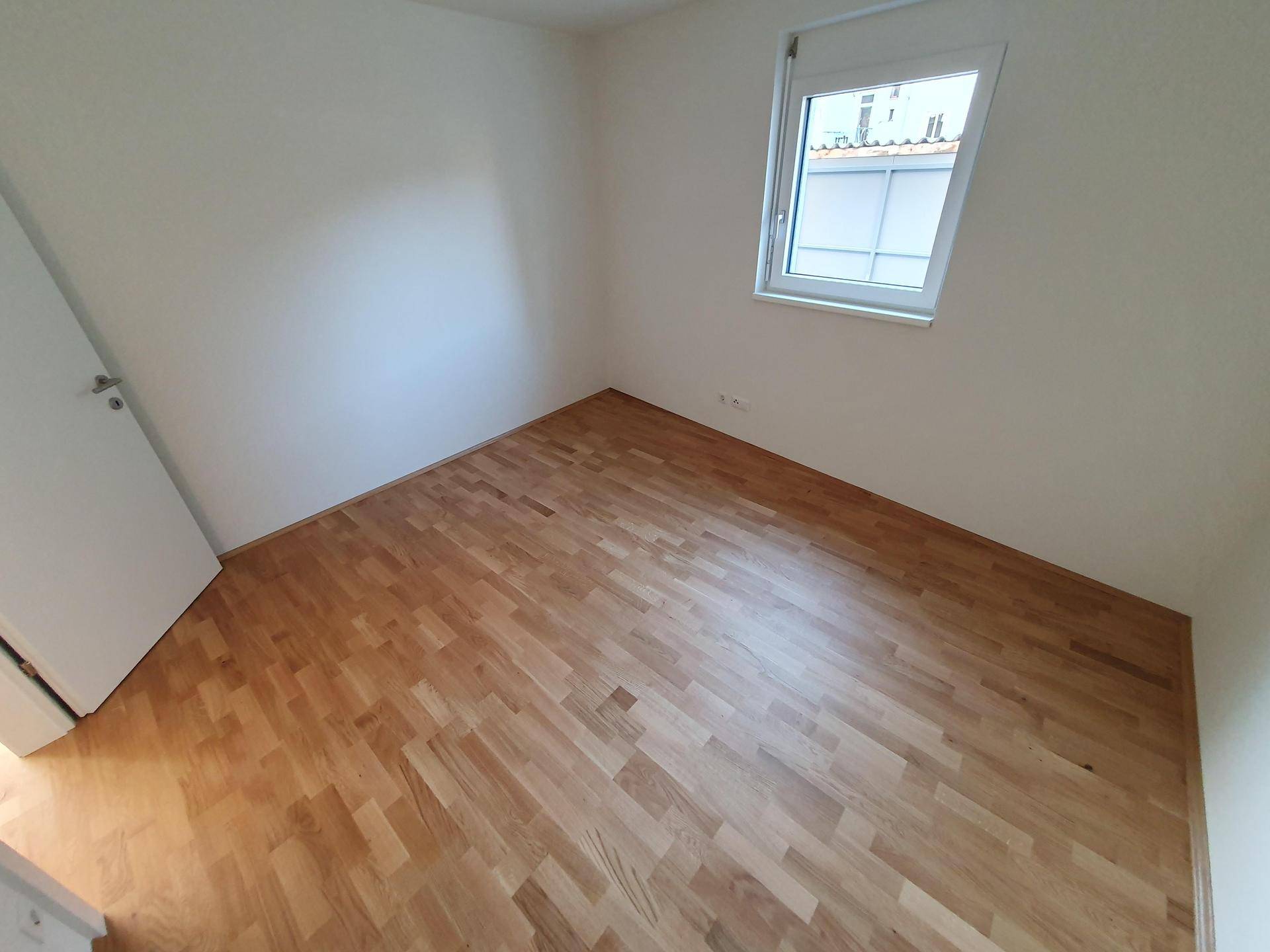 Helles Schlafzimmer mit Parkettboden und einem Fenster, bereit zur individuellen Gestaltung.