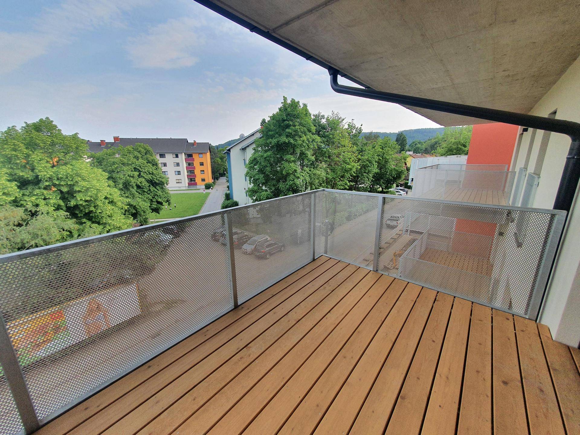 Geräumiger Balkon mit Holzboden und Metallgeländer, umgeben von Grünflächen.