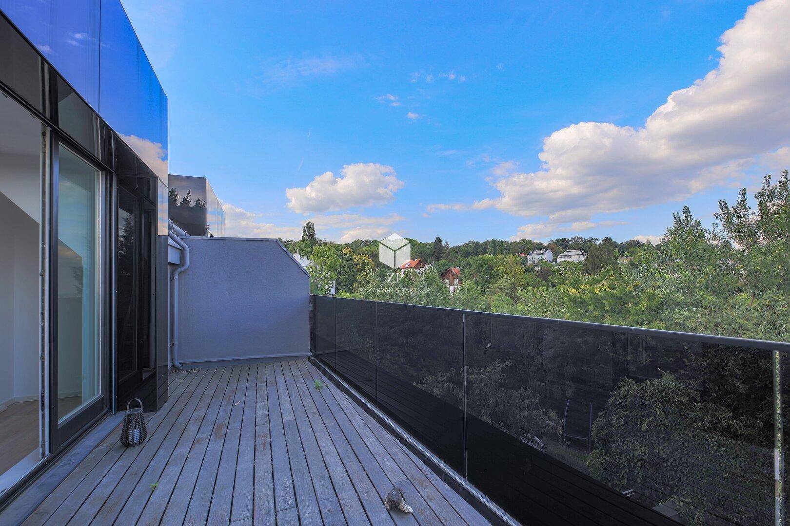 Moderner Balkon mit Holzboden und Glasgeländer, umgeben von viel Grün.