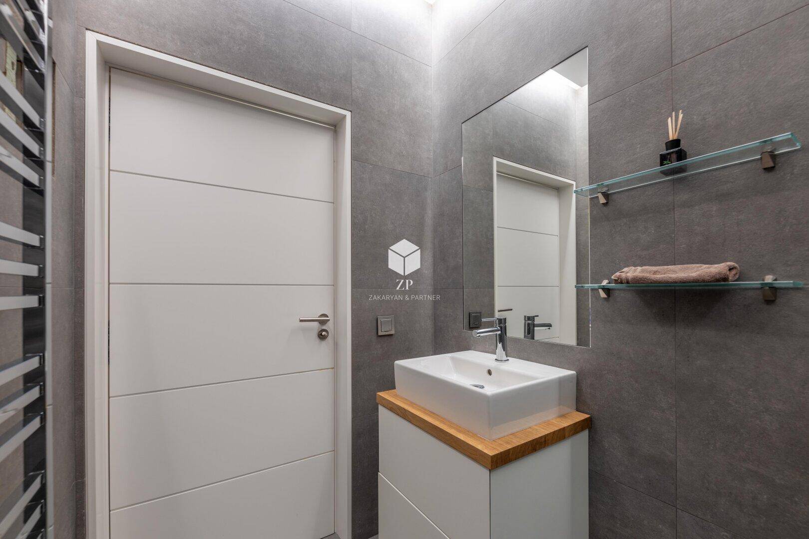Modernes Badezimmer mit Einzelwaschbecken, großem Spiegel und grauen Wandfliesen.