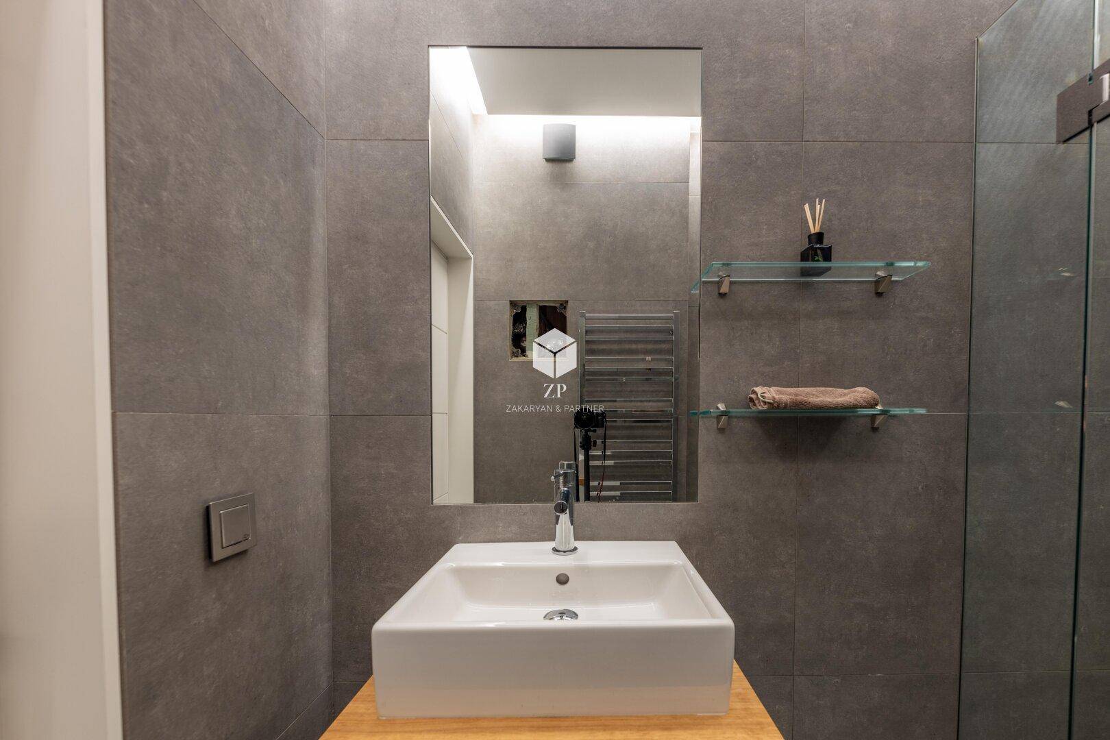 Stilvolles Badezimmer mit modernem Waschbecken, großem Spiegel und grauen Fliesen.