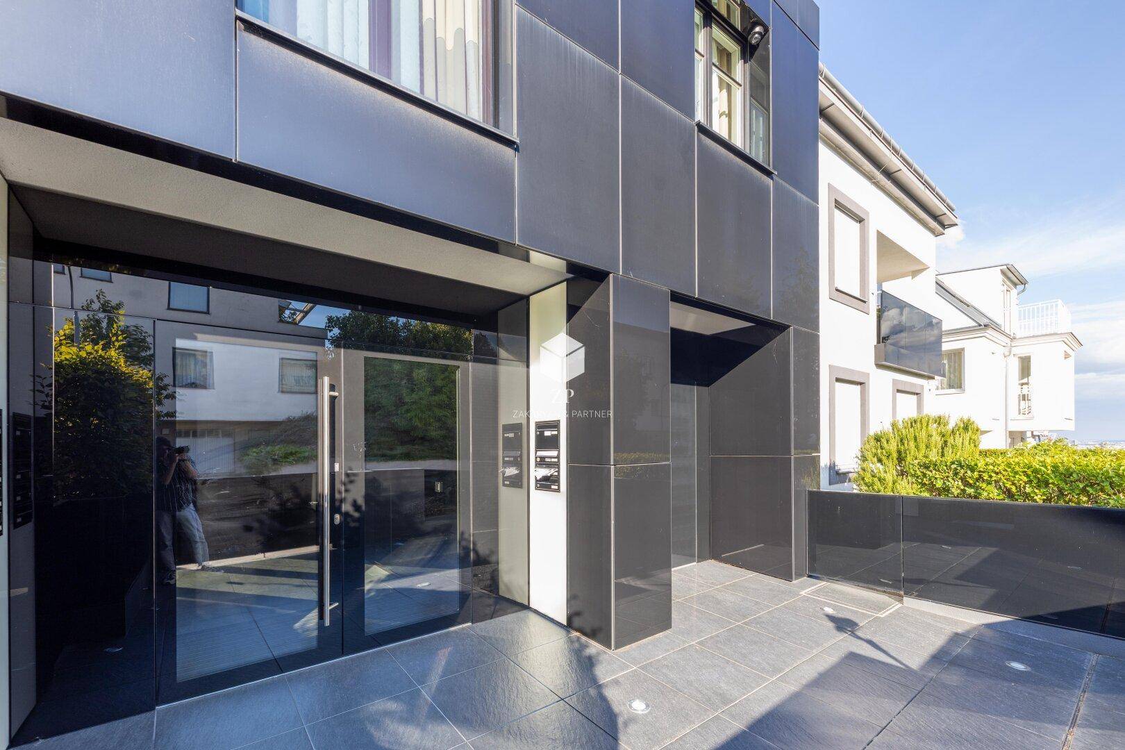 Eingangsbereich eines modernen Gebäudes mit dunkler Fassade und großen Glastüren.