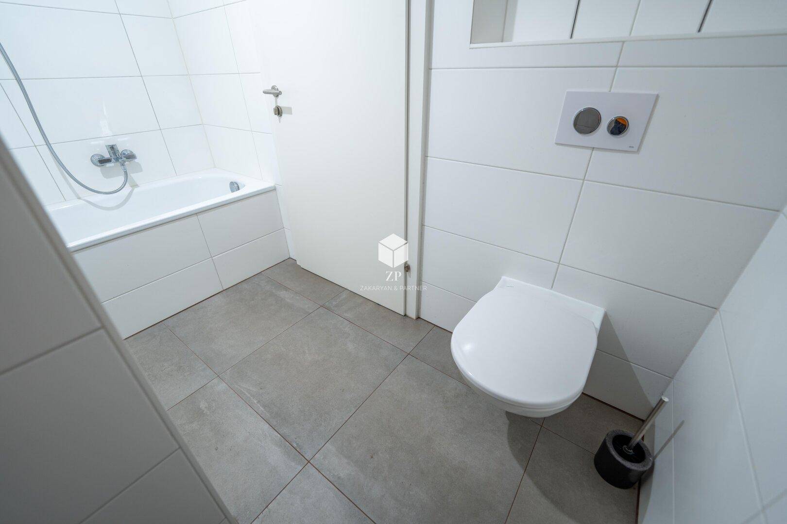 Helles Badezimmer mit Badewanne, WC und weißen Fliesen, ausgestattet für den täglichen Gebrauch.