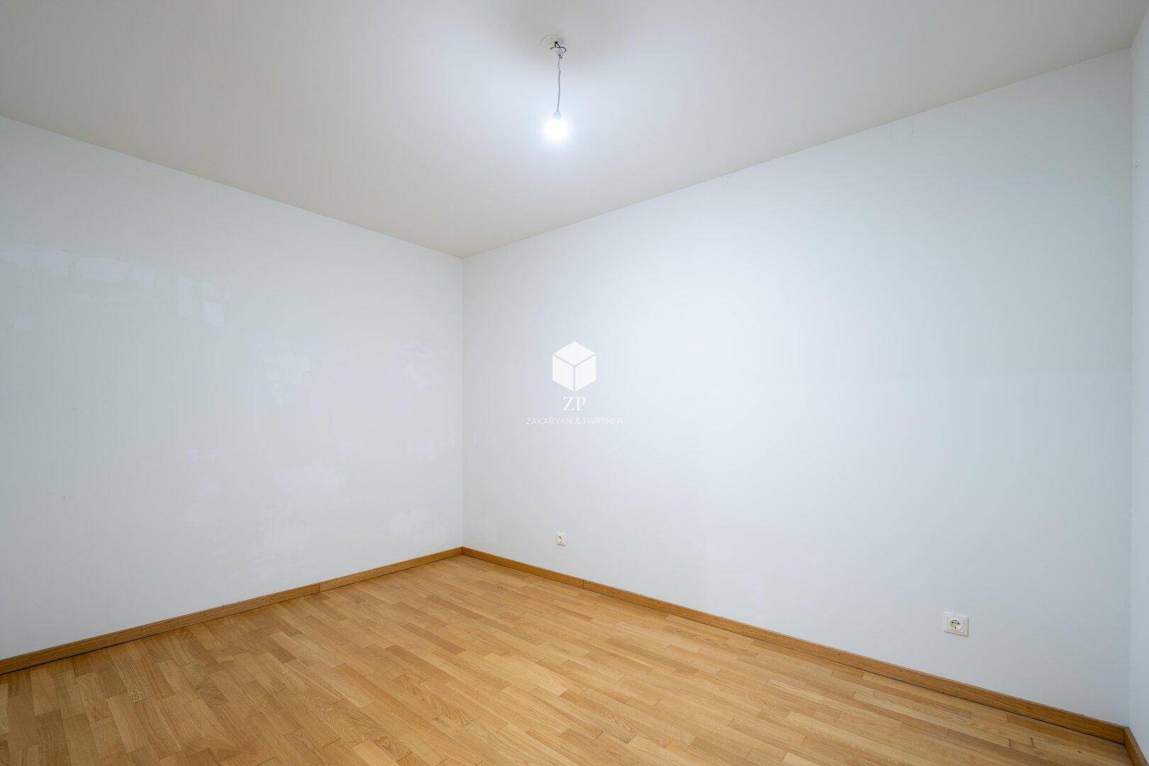 Leerer, heller Raum mit weiß gestrichenen Wänden und einem warmen Holzparkettboden.