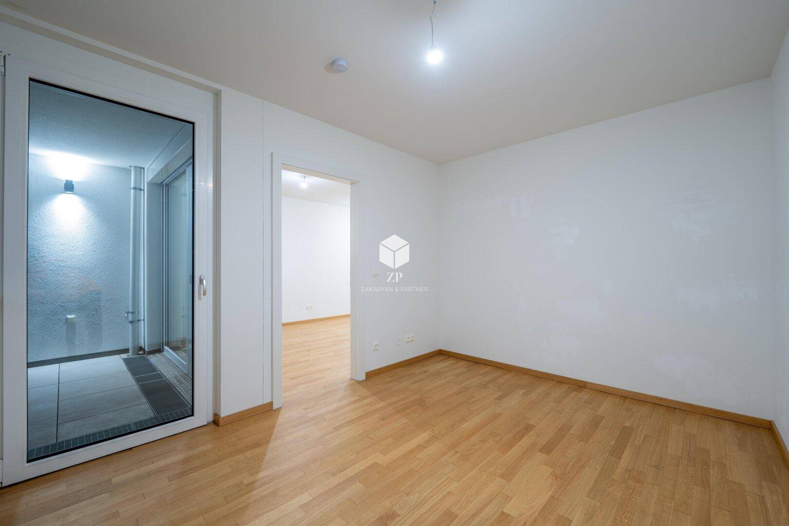 Heller Wohnraum mit Holzboden und Zugang zu einem Balkon oder einer Terrasse durch eine Glastür.