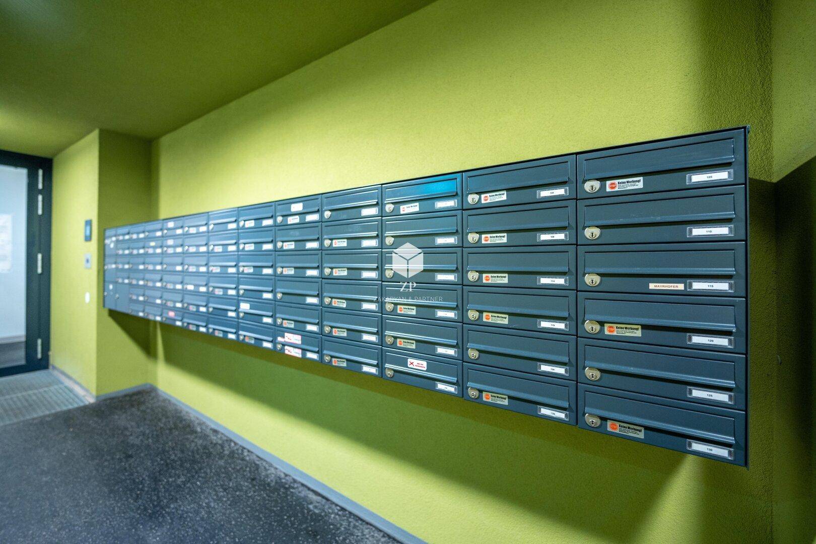 Lange Reihe von Briefkästen an einer leuchtend grünen Wand im Eingangsbereich eines Mehrfamilienhauses.
