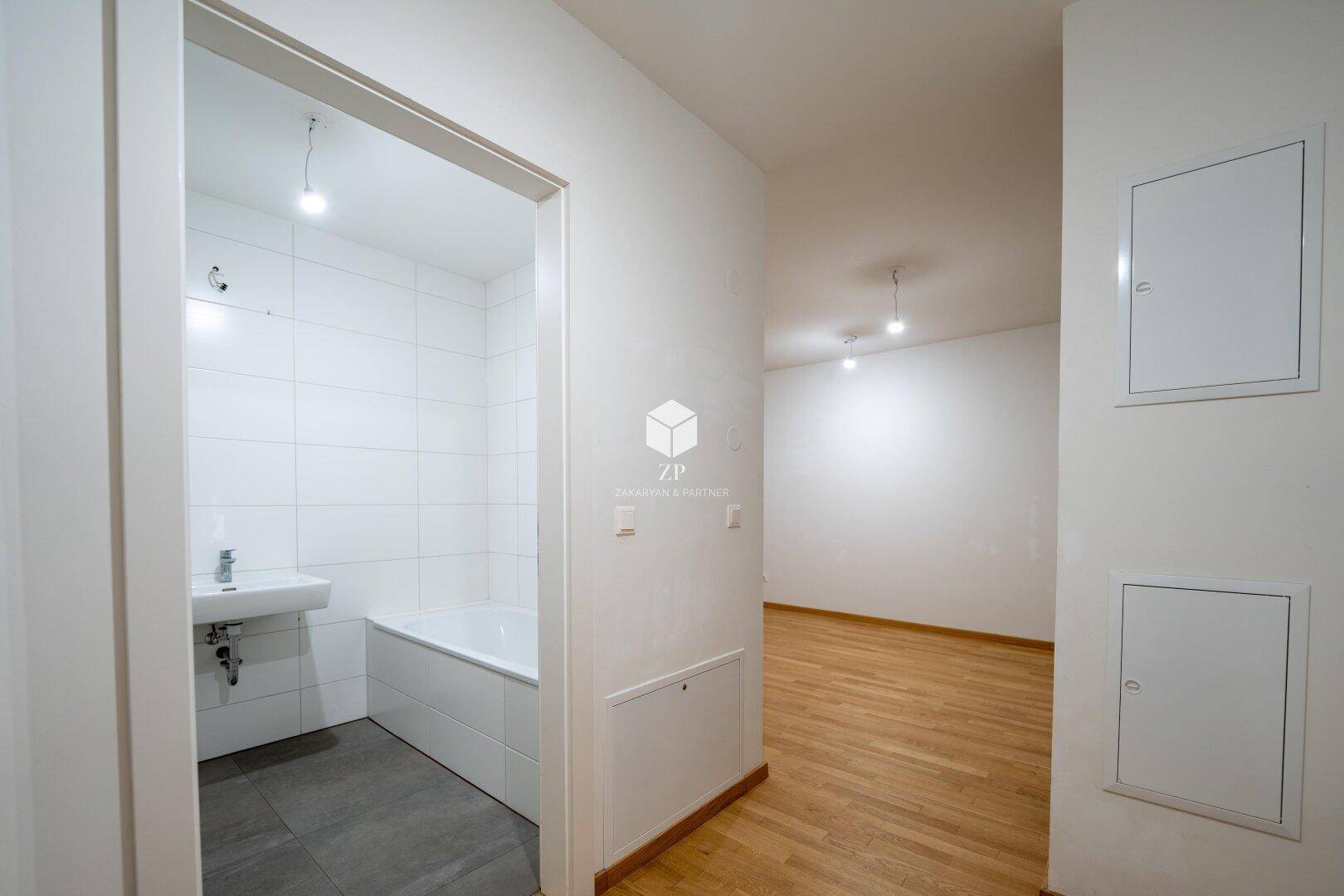 Heller Flur mit Holzboden und Zugang zu einem Badezimmer mit Badewanne und Waschbecken.