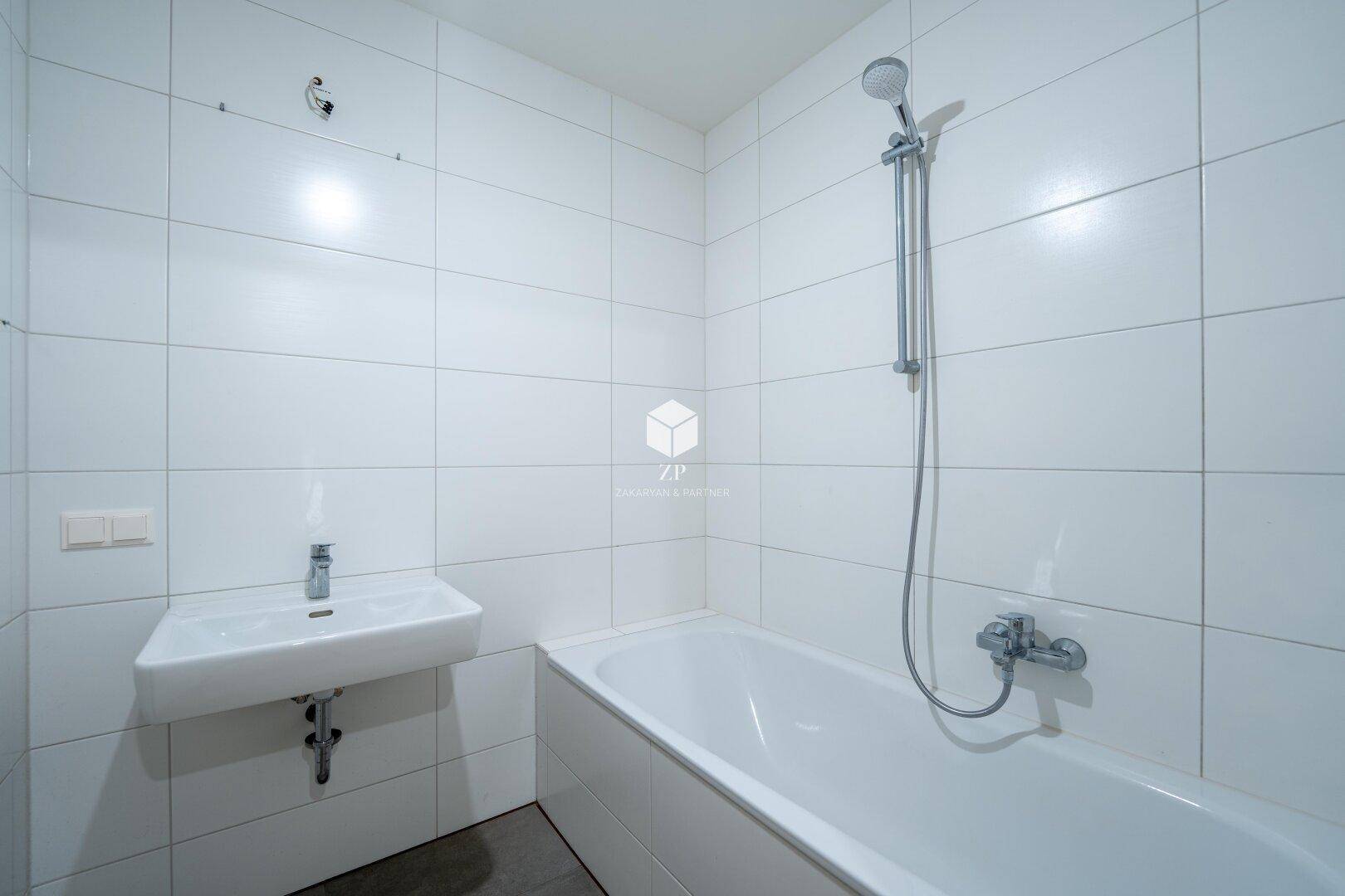 Modern gefliestes Badezimmer mit Badewanne, Duscharmatur und einem schlichten Waschbecken.
