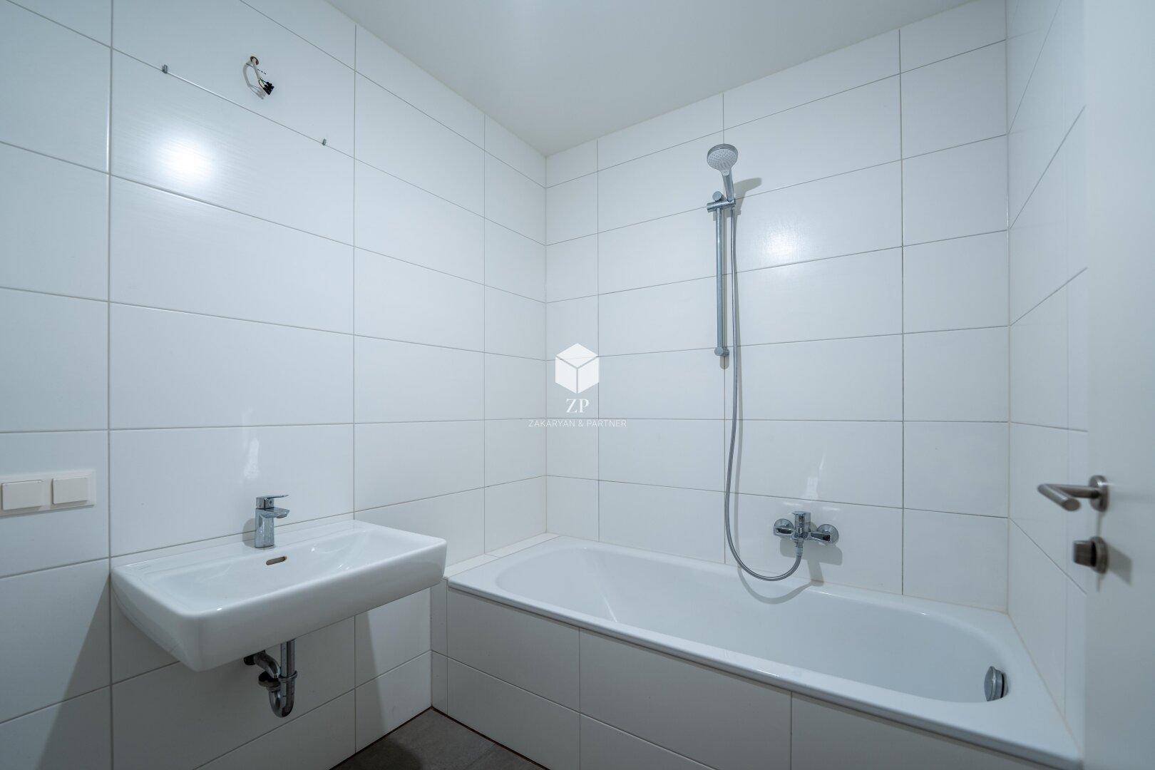 Sauberes Badezimmer mit weißen Wandfliesen, Badewanne mit Duschfunktion und Waschbecken.