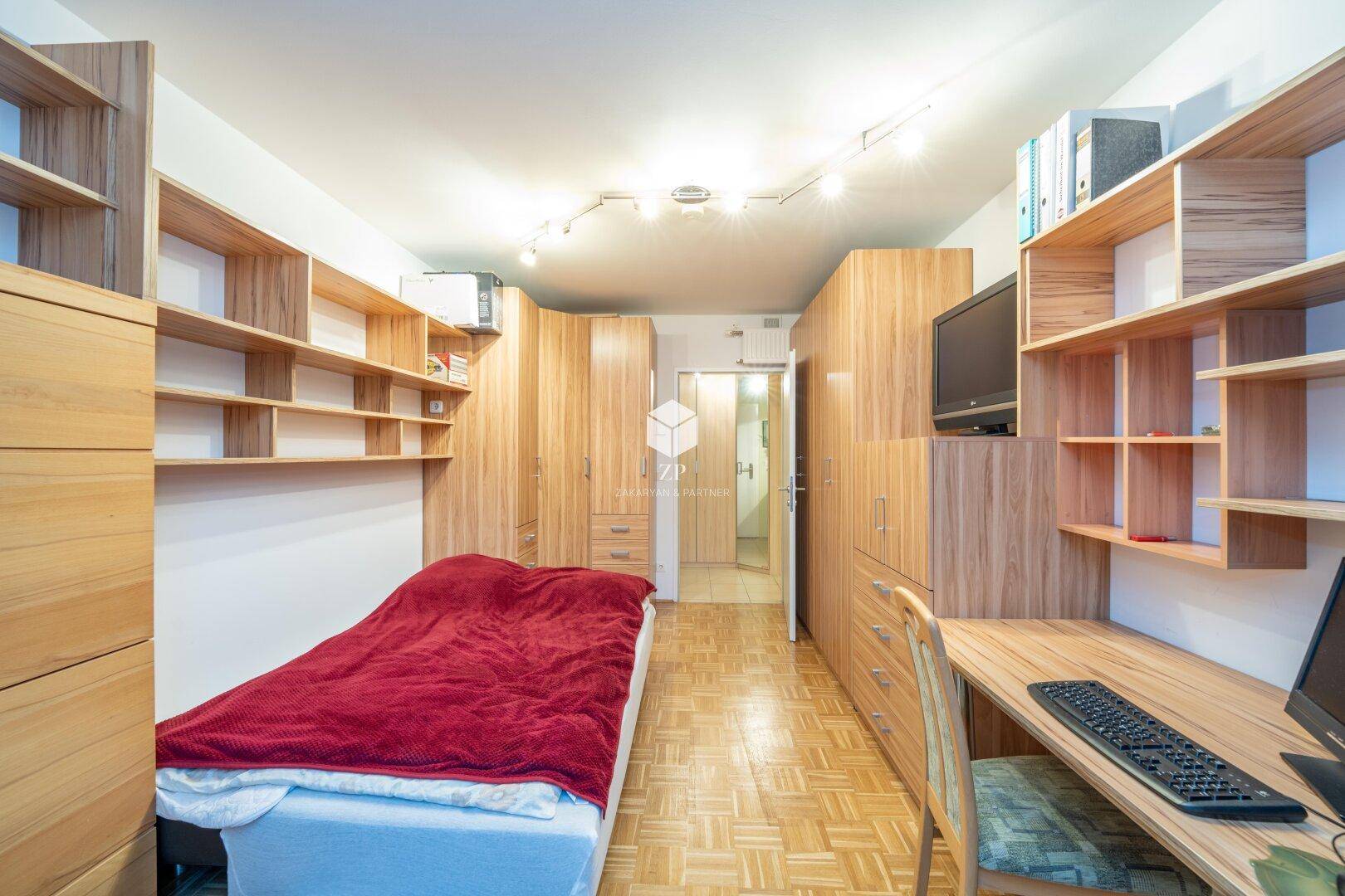 Schlafzimmer mit Einzelbett, Schreibtisch und umfangreichen Holzmöbeln.