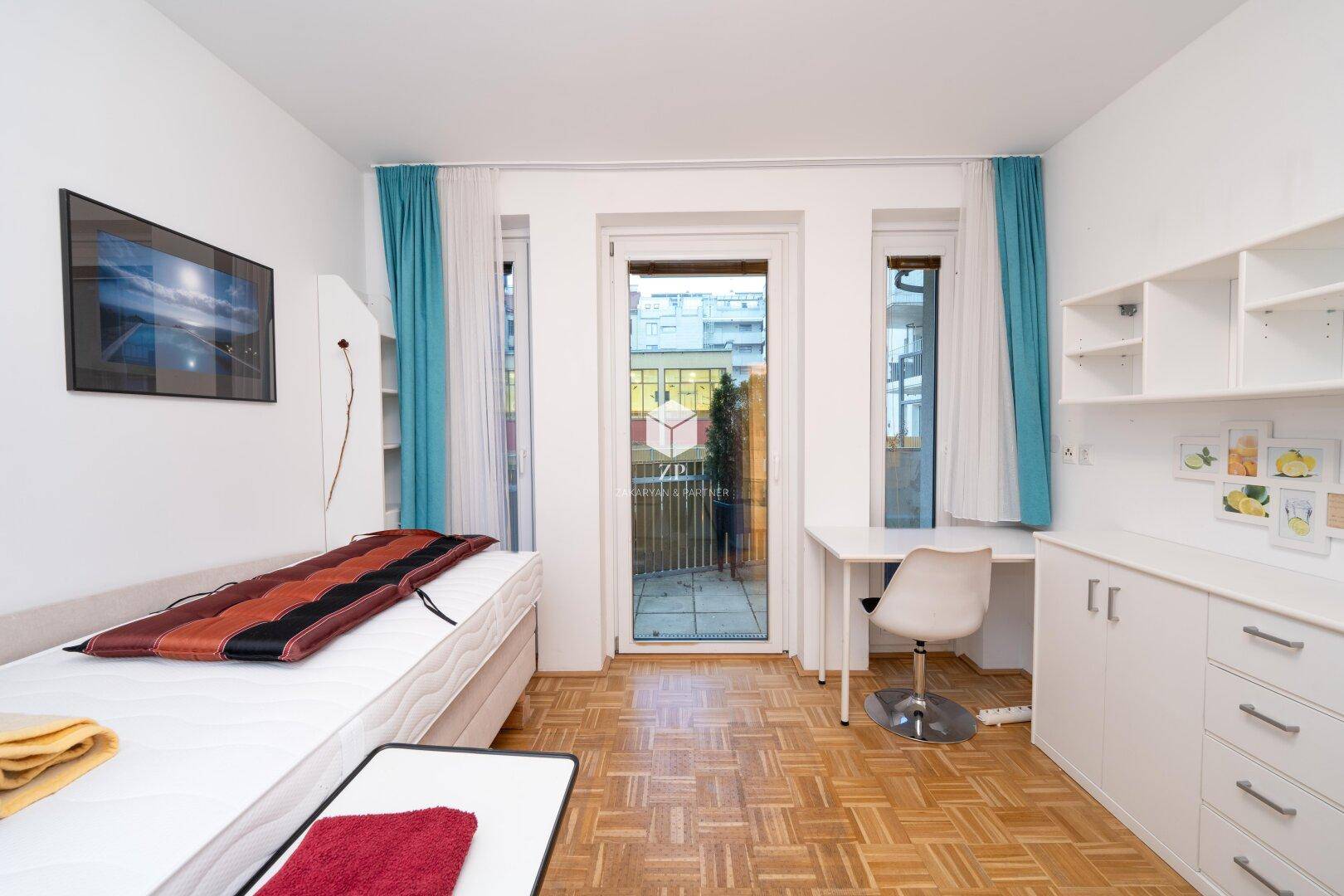 Helles Schlafzimmer mit Parkettboden, Schreibtisch und Zugang zum Balkon.