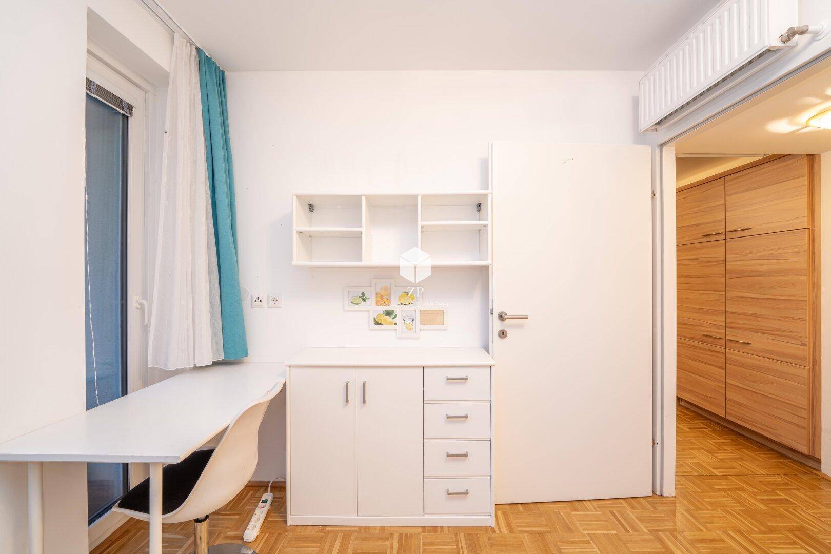 Arbeitsbereich im Schlafzimmer mit Schreibtisch, Regalen und weißem Kleiderschrank.