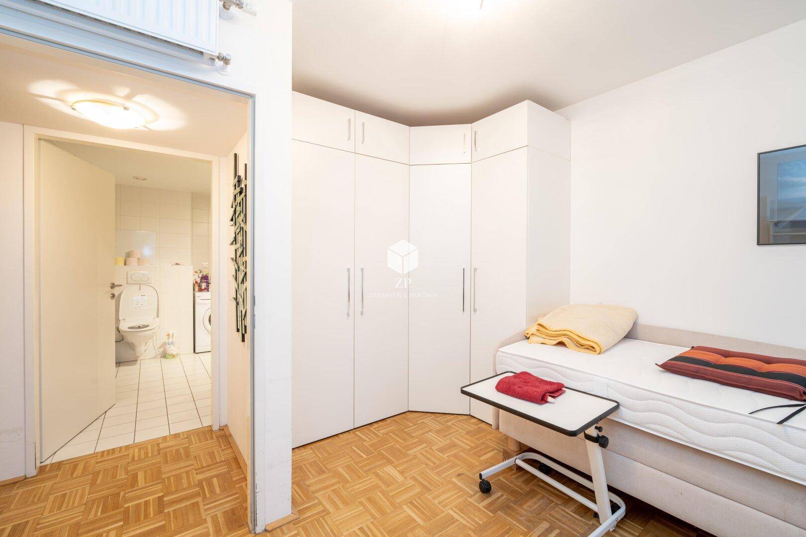 Schlafzimmer mit Einzelbett und weißem Eckkleiderschrank, angrenzend an das Badezimmer.