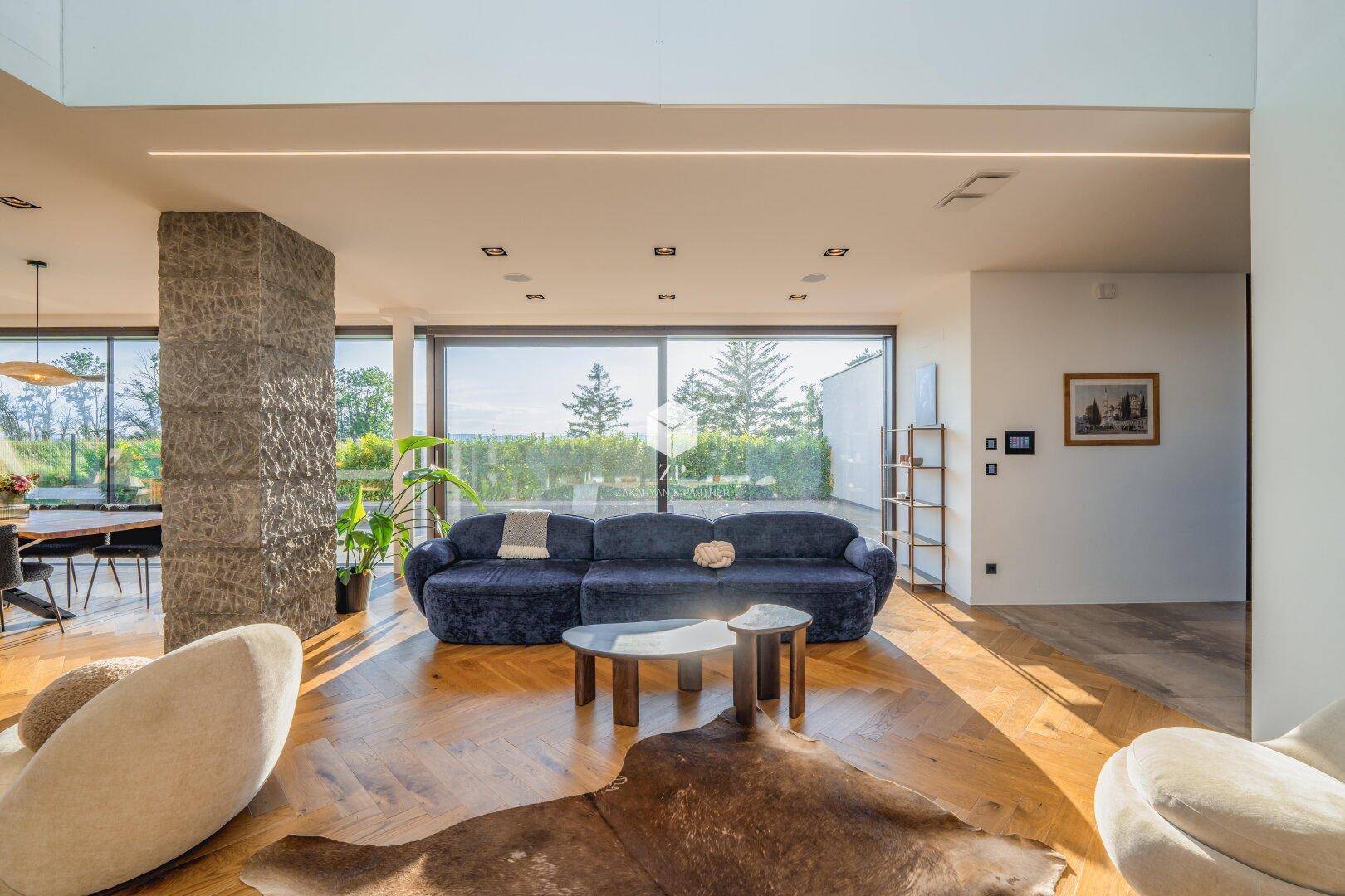Geräumiges Wohnzimmer mit blauer Couch, Steinsäule und großen Fenstern mit Blick in die Natur.