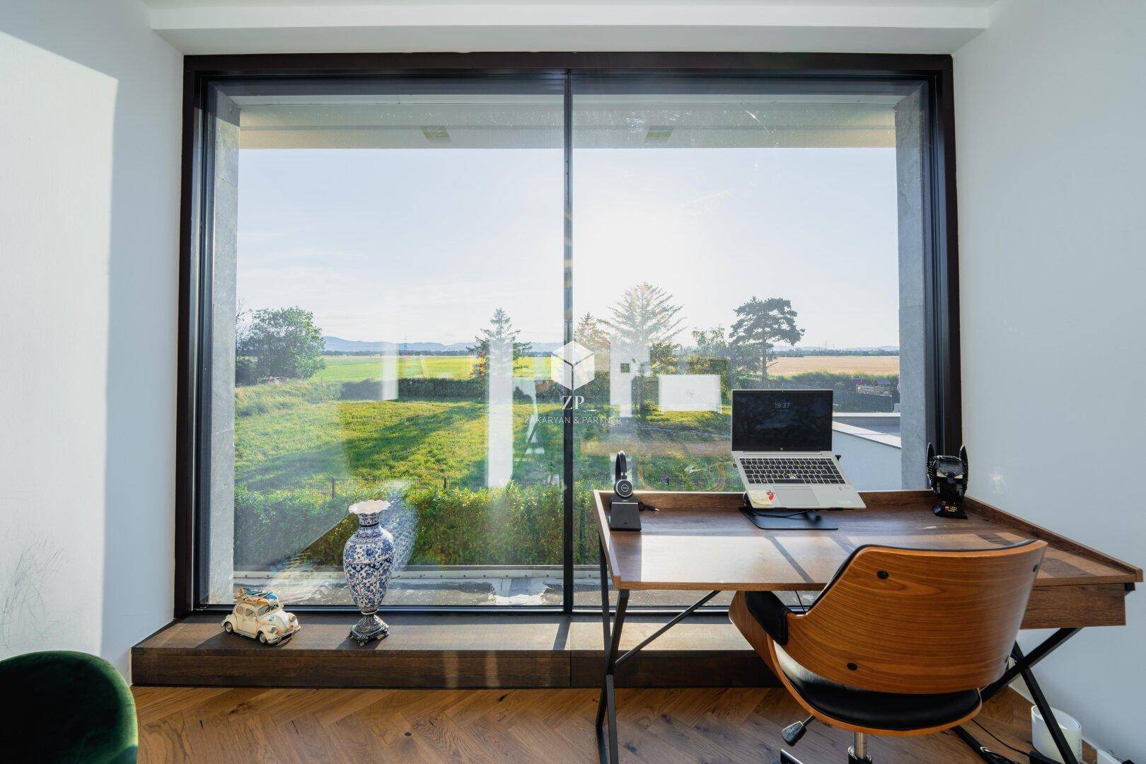 Helles Arbeitszimmer mit Schreibtisch und großem Fenster, das einen weiten Blick über die grüne Landschaft bietet.