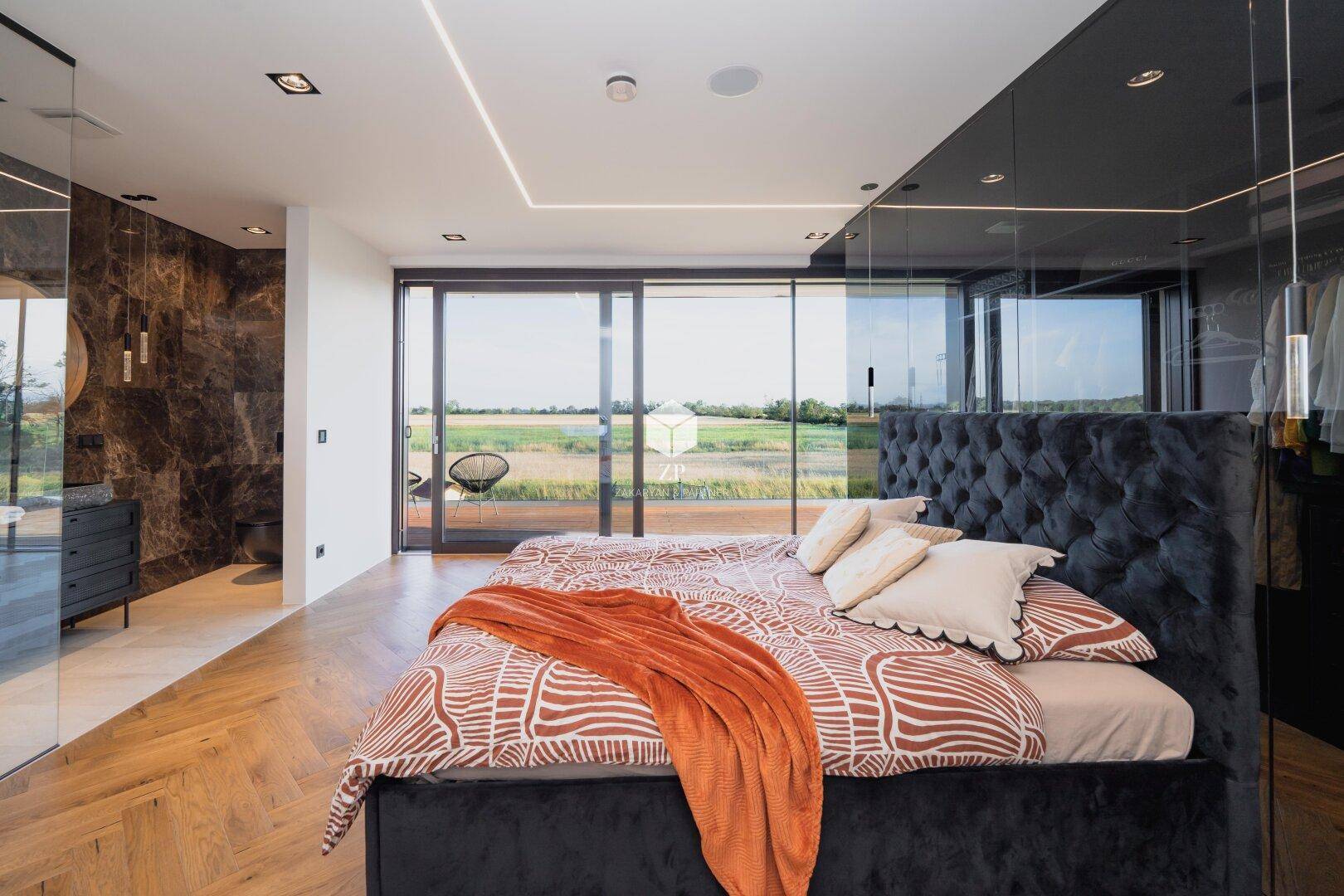 Elegantes Schlafzimmer mit großem Bett, Glaswand und Zugang zu einem Außenbereich mit weitem Blick.