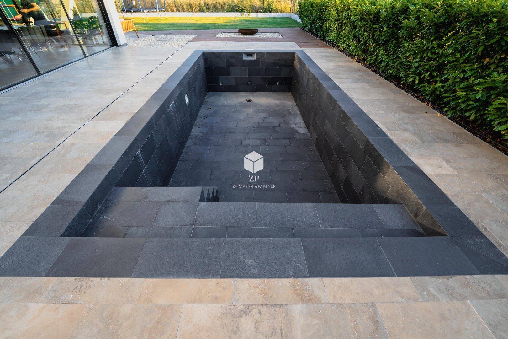 Rechteckiger Swimmingpool mit dunklen Fliesen, umgeben von einer gepflegten Terrasse und Hecke.