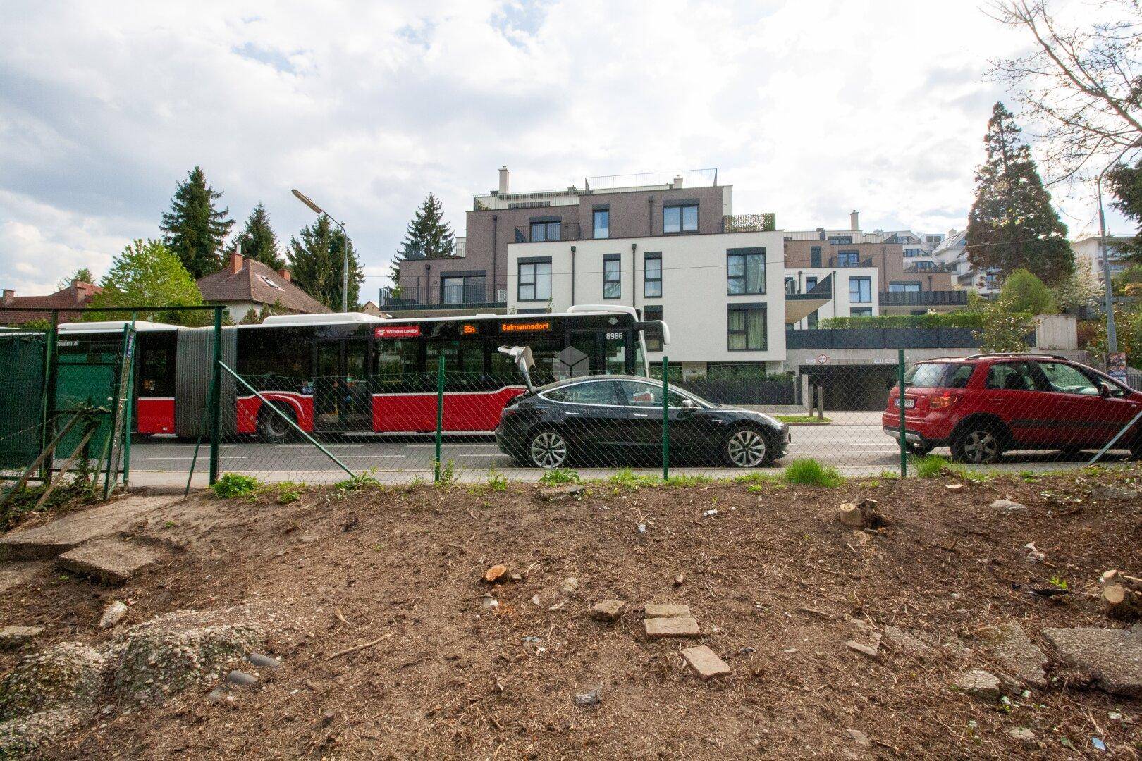 Blick vom unbebauten Grundstück auf eine Straße mit einem vorbeifahrenden Bus und parkenden Autos.