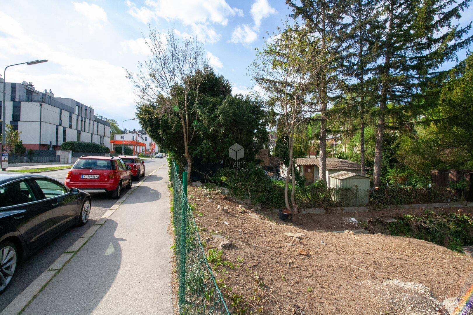 Unbebautes Grundstück an einer Straße mit parkenden Autos und üppiger Vegetation im Hintergrund.